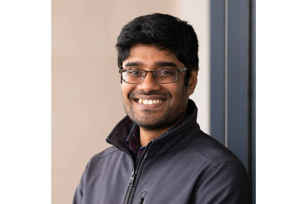 A man wearing glasses and a gray sweater smiles at a camera in front of a tan brown background.